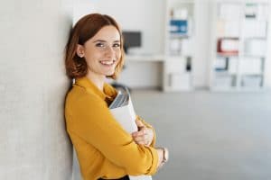 Happy young businesswoman clutching a large binder to her chest as she leans against an office wall grinning at the camera